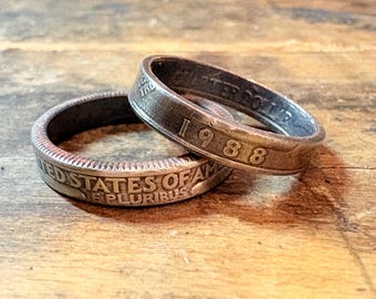 May include: Two handcrafted rings made from US coins. One ring displays the year "1988" and the other ring shows the words "UNITED STATES OF AMERICA". The rings are made of a silver-colored metal and rest on a wooden surface.