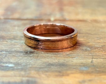 May include: A copper-colored metal ring with a smooth, polished finish and a flat band profile. The exterior surface of the ring features a subtle, engraved wheat stalk pattern that wraps around the circumference of the band.