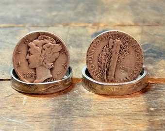 May include: Two coin rings, one with a profile and the text "IN GOD WE TRUST 1943", the other with a tree and the text "ONE DIME". Both rings are made of silver and sit on a wooden surface.