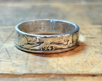 May include: A silver-toned coin ring crafted from a half-dollar coin. The exterior surface features embossed details including the year 1943 and the text HALF-DOLLAR. The metal has a polished finish with visible relief patterns from the original coin design.