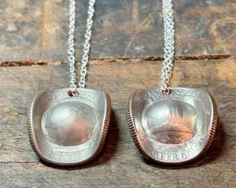 May include: Two silver-colored coin pendants, each shaped like a cowboy hat, hang from delicate silver chains. The pendants are made from quarters, with the coin details visible. The background is a rustic wooden surface.