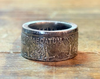 May include: A silver-toned metal ring crafted from a coin, featuring embossed details including a crown, swords, and the Latin text PRAEDATUM IN MUNDUM. The band has a wide, cylindrical shape with a weathered, metallic finish and visible relief.
