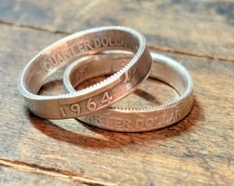 May include: Two silver rings made from 1964 quarter dollars. The rings are stacked on top of each other, with the text "QUARTER DOLLAR" and "1964" visible on the rings. The rings are on a wooden surface.