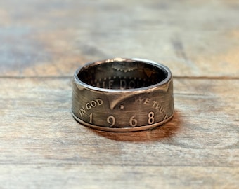 May include: A silver ring made from a 1968 half dollar coin. The ring features the words "IN GOD WE TRUST" and the year "1968" engraved on its surface. The ring is set against a wooden background.