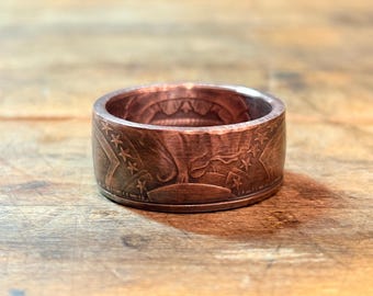 May include: A copper-colored ring with detailed engravings of stars and a stylized animal. The ring has a wide band and a textured surface, showcasing the intricate design. The ring is displayed on a wooden surface.