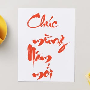 May include: A white card with red calligraphy, the words "Chúc Mừng Năm Mới" are written in a flowing script. A yellow teacup and saucer are on the left, and a yellow dish with a pen is on the right.