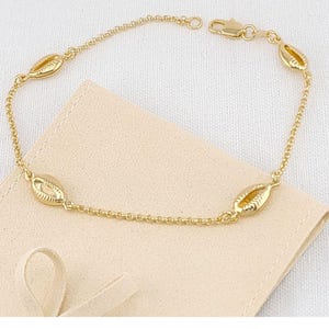 May include: A gold-colored bracelet featuring a delicate chain and several oval-shaped seashell charms. The bracelet is displayed on a light beige fabric, showcasing its intricate design and warm tones. The clasp is also gold.