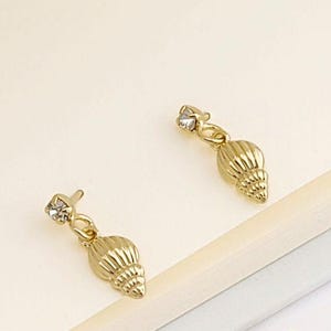 May include: Gold-colored seashell earrings with a small, clear gemstone at the top. The earrings have a textured, ridged design and are shown with two matching earring backs. The earrings are displayed on a light-colored surface.