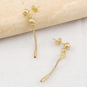May include: A pair of gold-colored dangle earrings. Each earring features a small ball stud, a textured cylindrical element, a delicate chain, and a small, hollow tube at the end. The earrings are displayed on a light-colored surface.