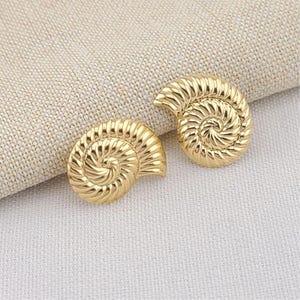 May include: Two gold-colored, seashell-shaped earrings. Each earring features a spiral design with detailed ridges, giving a textured appearance. The earrings are displayed on a neutral-colored fabric background, highlighting their metallic sheen.