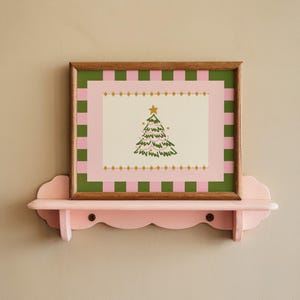 Puede incluir: Una impresión enmarcada de un árbol de Navidad con un borde a cuadros rosa y verde, exhibida en un estante rosa con volantes. La impresión presenta un árbol de Navidad verde con una estrella dorada sobre un fondo crema.