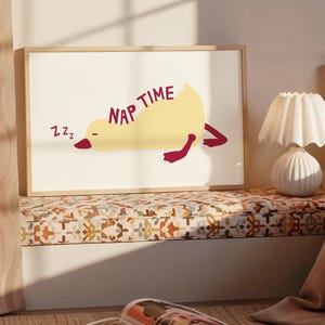 Puede incluir: Un cuadro enmarcado con un pato de dibujos animados amarillo en posición de dormir con las palabras "NAP TIME" en granate. El pato tiene pies y pico granates. El fondo es blanquecino.