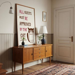 Puede incluir: Un aparador de madera de estilo moderno de mediados de siglo con tres cajones y dos puertas. Un póster enmarcado con el texto "ALL GUESTS MUST BE APPROVED BY THE CAT" y una ilustración de un gato gruñón está apoyado contra el aparador. Una lámpara y objetos decorativos están encima.