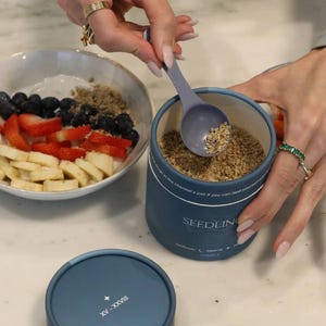 Puede incluir: Un recipiente azul con la etiqueta "SEEDLING" se está llenando con una cuchara. El recipiente está junto a un cuenco de fruta, que incluye fresas, arándanos y plátanos. La tapa del recipiente tiene los números romanos XV-XXVIII.