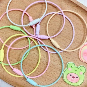 May include: A collection of colorful, pastel-colored keychains in shades of pink, green, yellow, and blue. Each keychain is a circular loop with a matching clasp. The keychains are arranged on a wooden surface, with a few decorative stickers.