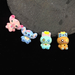 Puede incluir: Cuatro figuras de personajes de dibujos animados coloridos. Las figuras incluyen un conejito rosa, un extraterrestre azul, una muñeca verde azulado y una muñeca naranja. Cada figura tiene detalles únicos, incluyendo ojos de botón y costuras. Las figuras están sobre una superficie oscura.