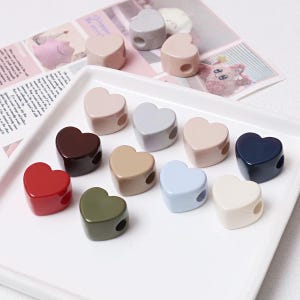 May include: A collection of heart-shaped beads in various solid colors including red, dark brown, olive green, light blue, cream, navy blue, grey, and beige, arranged on a white square tray with a smooth, glossy finish and a side hole.