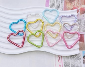 May include: A collection of heart-shaped carabiner clips in various colors, including blue, red, yellow, green, pink, and purple. The clips are made of metal and have a spring-loaded gate. The image also includes text from a printed page.