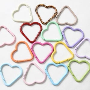 May include: A collection of heart-shaped carabiner clips in various colors, including red, pink, orange, yellow, green, blue, and purple. One clip features a leopard print design. The clips are arranged on a white surface.