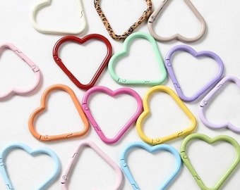 May include: A collection of heart-shaped carabiner clips in various colors, including red, pink, orange, yellow, green, blue, and purple. One clip features a leopard print design. The clips are arranged on a white surface.