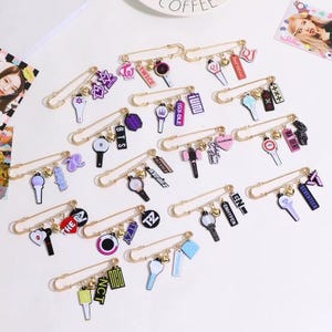 May include: A collection of gold-colored safety pins, each adorned with various charms. The charms include miniature logos, musical instruments, and text, such as "TWICE", "BTS", and "SEVENTEEN". The pins are arranged on a white surface, suggesting a craft or accessory theme.