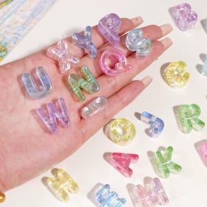 Puede incluir: Una colección de abalorios de letras del alfabeto translúcidas y coloridas en varios colores, incluyendo rosa, verde, azul y amarillo. Las letras están dispersas sobre una superficie, algunas sujetas en una mano. Las letras son de un material transparente e iridiscente.