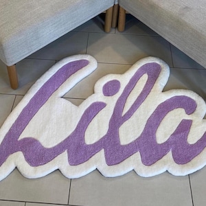 May include: A custom tufted rug featuring the name Lila written in a cursive script. The lettering is rendered in a soft lavender purple yarn, set against a thick, plush white background that follows the contour of the letters.