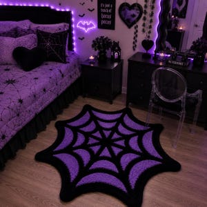 Puede incluir: Una alfombra circular con un patrón de telaraña en negro y morado. La alfombra tiene una superficie texturizada y con mechones, con un diseño de red distintivo que irradia desde el centro, alternando secciones moradas y negras.