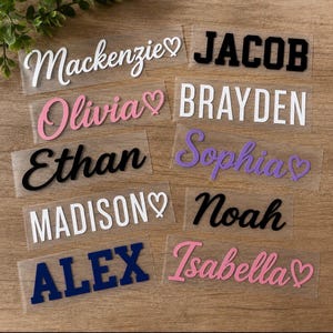 May include: A collection of vinyl name decals in various fonts and colors, including Mackenzie, Jacob, Olivia, Brayden, Ethan, Sophia, Madison, Noah, Alex, and Isabella. Several names feature a small heart icon, presented on clear backing sheets.