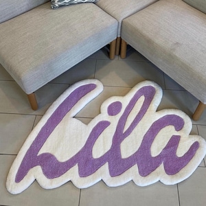 May include: A custom tufted rug featuring the name Lila in a cursive script. The lettering is rendered in a soft purple yarn, outlined by a thick border of white yarn, creating a plush, textured surface with a distinct, irregular shape.