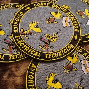 Könnte beinhalten: Mehrere gestickte Aufnäher mit einer gelben Cartoonfigur und dem Text "Coast Guard Aviation Avionics Electrical Technician". Die Aufnäher haben ein kreisförmiges Design mit schwarzem Rand und sind grau und gelb.