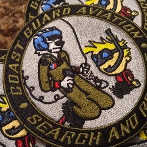 Puede incluir: Parches bordados con un diseño de Búsqueda y Rescate de Aviación de la Guardia Costera. Los parches circulares presentan una ilustración de dibujos animados de un personaje con traje de vuelo y otro con un esnórquel. Texto: 'Coast Guard Aviation Search and Rescue'.