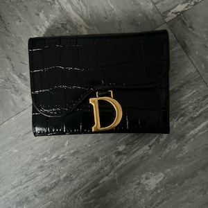 May include: A black wallet with a glossy crocodile-embossed texture featuring a curved flap closure. A prominent, gold-toned metal letter D is centred on the front, serving as a decorative accent on the flap. The wallet has a rectangular shape.
