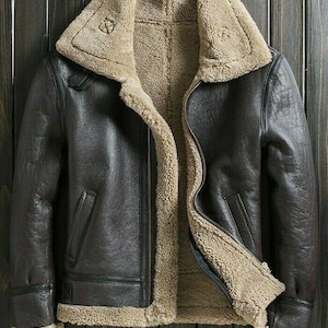 Puede incluir: Chaqueta de cuero marrón con forro y cuello de borrego beige. La chaqueta tiene cierre de cremallera, dos bolsillos delanteros y un estilo aviador clásico. La chaqueta se muestra sobre un fondo de madera oscura.