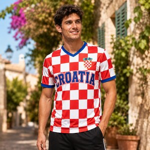 Pode incluir: Uma t-shirt de manga curta com decote em V, apresentando um padr&atilde;o de xadrez vermelho e branco em toda a pe&ccedil;a. A palavra CROATIA est&aacute; impressa em letras azuis grandes no peito, com um pequeno emblema do bras&atilde;o croata acima do texto.