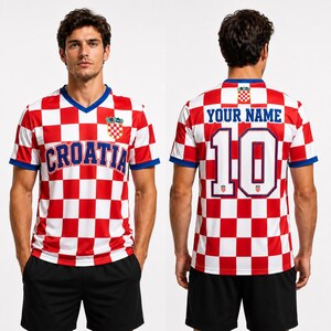 Pode incluir: Uma camisola de futebol aos quadrados vermelhos e brancos com decote em V e acabamentos azuis. A frente apresenta o texto CROATIA e um emblema. As costas exibem o texto YOUR NAME acima do n&uacute;mero 10, com um pequeno emblema na gola e no n&uacute;mero.