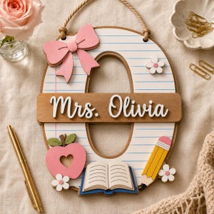 May include: A wooden letter O wall hanging with a notebook paper pattern, featuring a pink bow, a pink apple with a heart cutout, an open book, and a yellow pencil. A horizontal wooden plaque across the center displays the white text Mrs. Olivia.