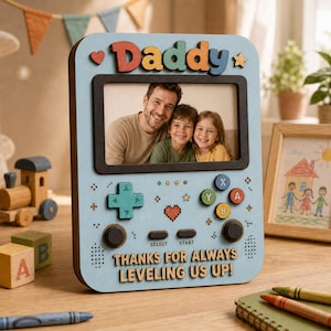 May include: A light blue wooden photo frame in the shape of a video game controller, with the word "Daddy" in colorful letters at the top. The frame holds a family photo and includes the text "THANKS FOR ALWAYS LEVELING US UP!"
