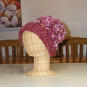 May include: A burgundy and pink knitted hat with a fluffy texture. The hat is displayed on a wicker mannequin head.