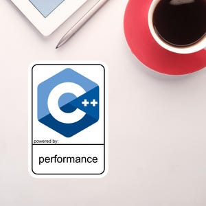 May include: A sticker with the C++ programming language logo, a blue hexagon with a white "C" and "++" symbols. The text "powered by: performance" is below the logo. A cup of coffee and a tablet are also visible.