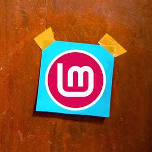 May include: A blue square sticker with a white and magenta logo, taped to a wooden surface with two pieces of yellow tape. The logo features the letters "lm" in white within a magenta circle.