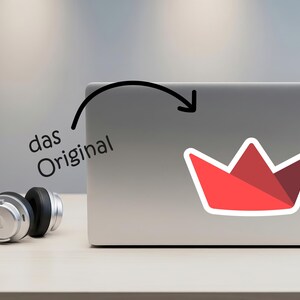 Op de afbeelding: Een rode sticker in de vorm van een kroon met een witte rand zit op een laptop. De sticker heeft een geometrisch ontwerp. De tekst "das Original" staat boven de laptop. Een zilveren koptelefoon ligt op de tafel.