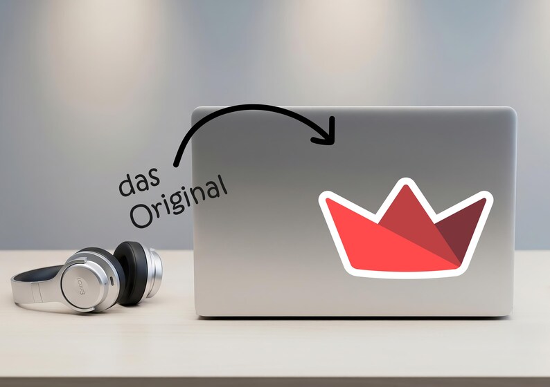 Op de afbeelding: Een rode sticker in de vorm van een kroon met een witte rand zit op een laptop. De sticker heeft een geometrisch ontwerp. De tekst "das Original" staat boven de laptop. Een zilveren koptelefoon ligt op de tafel.