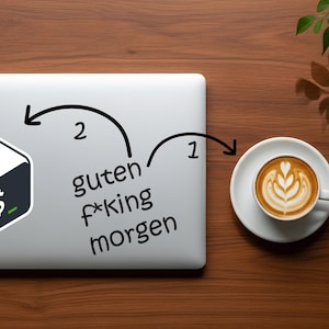 May include: A laptop with a sticker featuring a stylized terminal icon and the text "guten f*king morgen". A cup of coffee with latte art sits on a saucer to the right. The items are on a wooden surface.