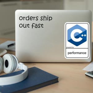 May include: A laptop with a sticker that says "orders ship out fast." A C++ logo sticker is also on the laptop. White headphones, a blue notebook, a cup, and a calculator are on the wooden desk.