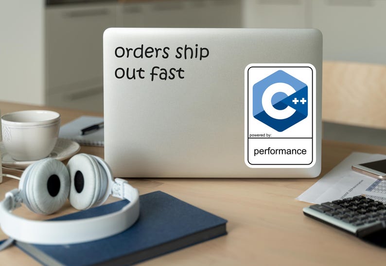 May include: A laptop with a sticker that says "orders ship out fast." A C++ logo sticker is also on the laptop. White headphones, a blue notebook, a cup, and a calculator are on the wooden desk.