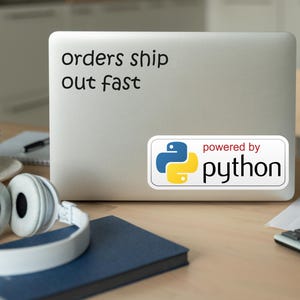 May include: A laptop with the text "orders ship out fast" and a "powered by python" sticker. White headphones, a blue notebook, a cup, and a calculator are on a wooden desk.