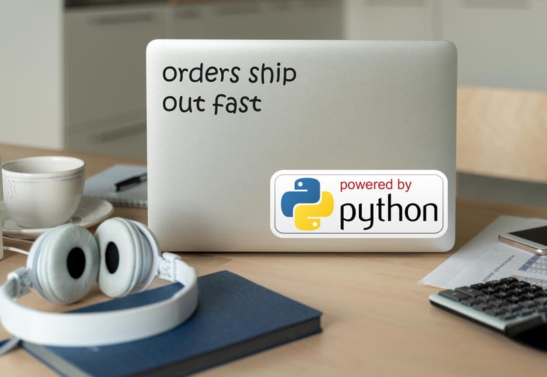 May include: A laptop with the text "orders ship out fast" and a "powered by python" sticker. White headphones, a blue notebook, a cup, and a calculator are on a wooden desk.