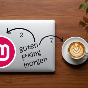 May include: A laptop with a pink and white logo sticker and the German phrase "guten f*king morgen" is next to a cup of coffee with latte art on a wooden surface.
