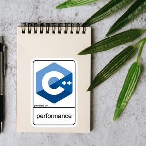 May include: A notepad with a spiral binding, a silver pen, and a sticker featuring a blue and white C++ logo with the text "performance". Green leaves are in the background. The items are on a grey surface.